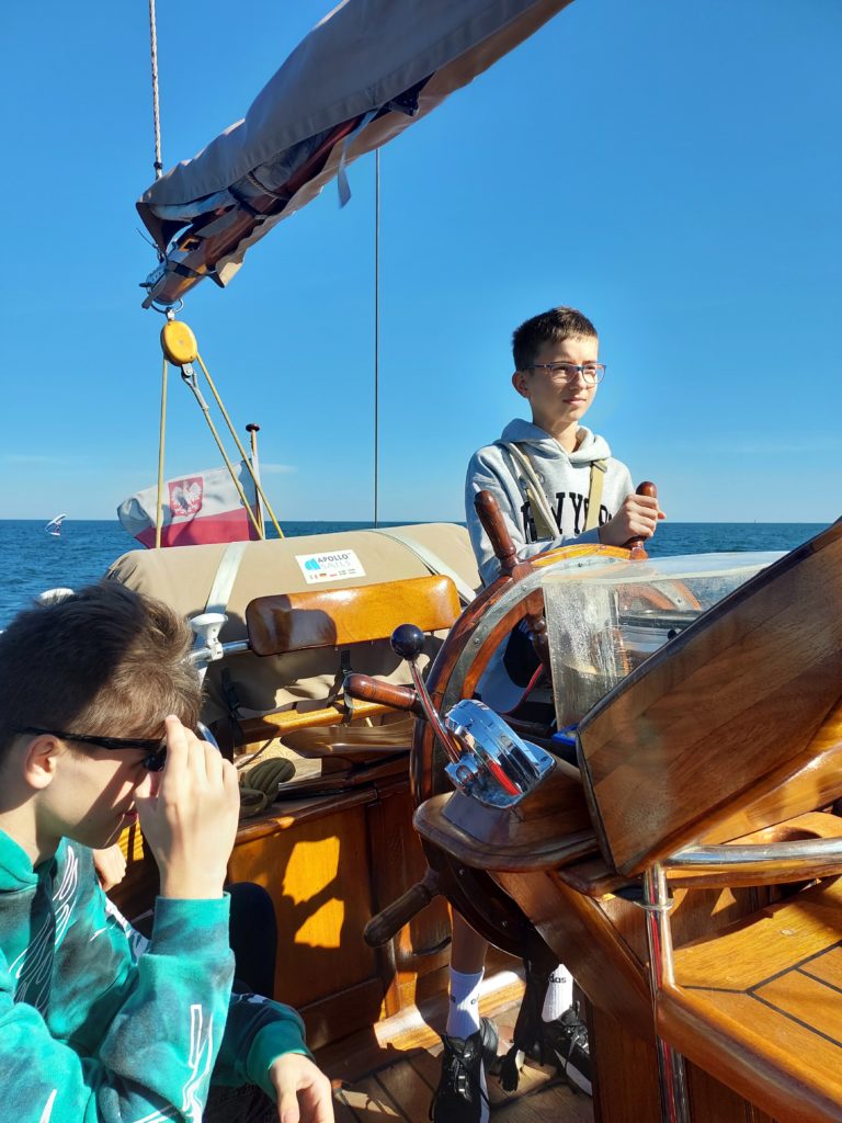 Chłopiec w okularach stoi na statku i trzyma koło sterowe. Obok siedzi chłopiec w zielonej bluzie i trzyma rękę na okularach przeciwsłonecznych. W tle jest flaga Polski i morze.