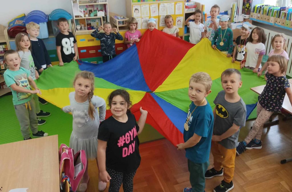 Zdjęcie przedstawia grupę dzieci, które bawią się wspólnie przy użyciu chusty animacyjnej.