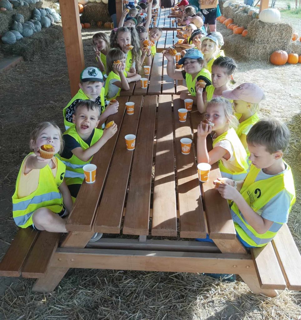 Grupa ok. 20 dzieci siedzi przy stolikach. Piją herbatkę i jedzą dyniowe babeczki.