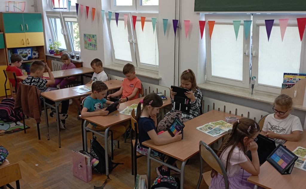 W sali lekcyjnej przy pięciu stolikach w rzędzie pod oknem siedzi 10 uczniów klasy pierwszej. Każdy uczeń rozwiązuje quiz na szkolnym iPadzie. W tle kolorowe szafki