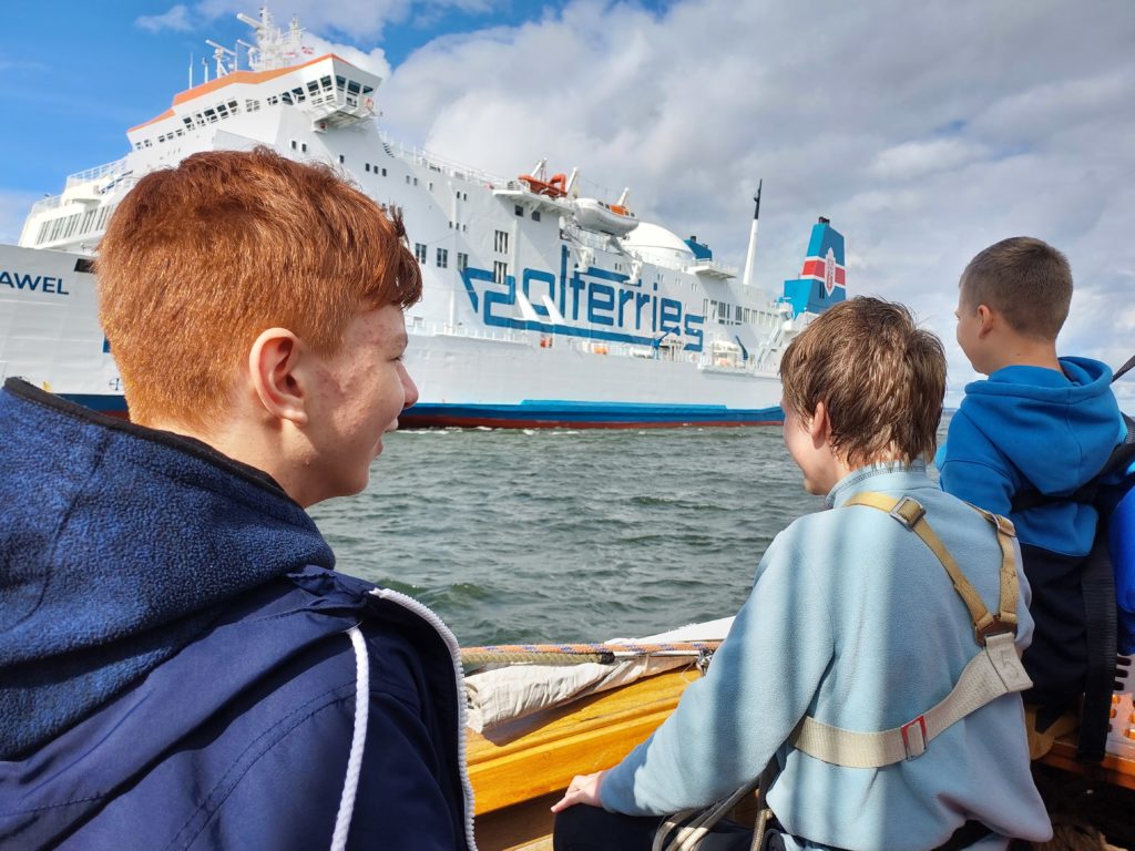 Trzech chłopców stoi na statku tyłem. Przed nimi jest na wodzie duży prom pasażerski.
