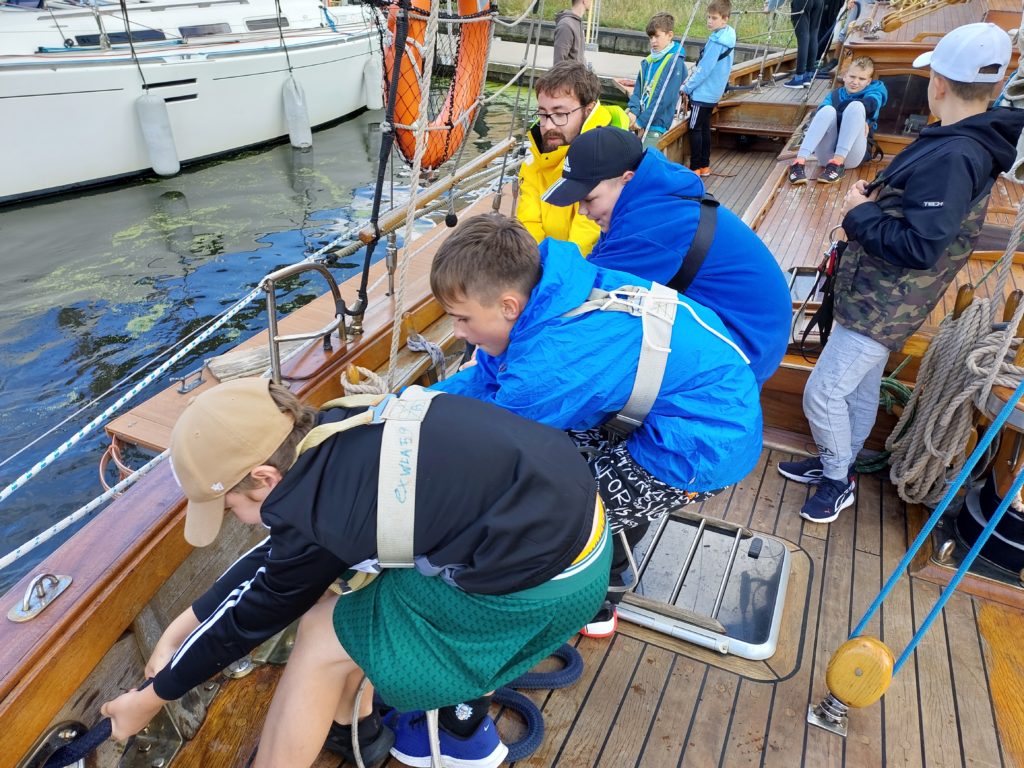 Czterech chłopców ciągnie linę na statku. W tle na drewnianym pokładzie stoją inni chłopcy.