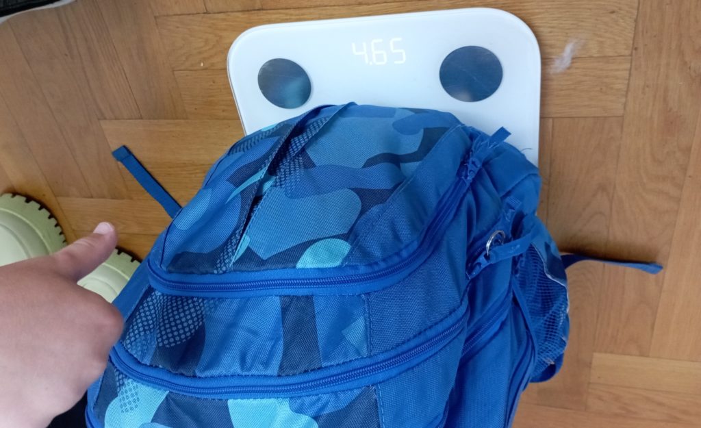 Na podłodze stoi elektroniczna waga ze szkolnym plecakiem. Wyświetlacz wagi wskazuje, że plecak waży 4,65 kg. Dziecięca dłoń wskazuje opuszczony w dół kciuk