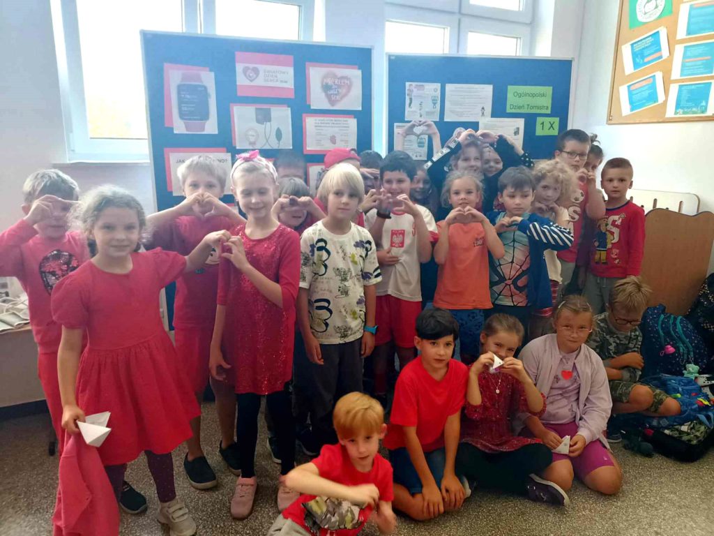24 uczniów klasy 2a pozuje do zdjęcia w czerwonych strojach na tle tablic z informacjami na temat Światowego Dnia Serca.