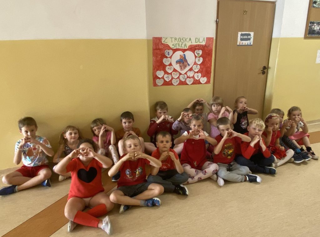 Na szkolnym korytarzu na tle wiszącego na ścianie plakatu z sercami siedzą uczniowie klasy 0b w czerwonych strojach. Dłonie mają ułożone w kształcie małych serc.
