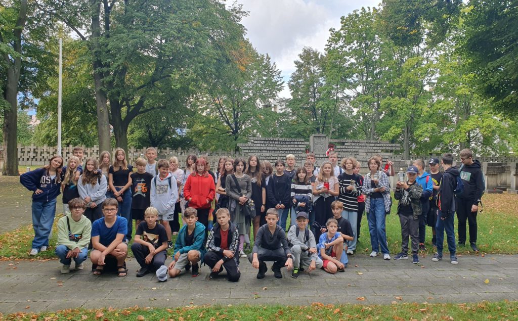 Uczniowie stoją w grupie na tle Pomnika ku czci kolejarzy i celników z Szymankowa. W oddali zieleń drzew