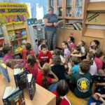 Dzieci siedzą na podłodze w bibliotece w siadzie skrzyżnym. Patrzą na stojącą przed nimi bibliotekarkę czytającą im tekst. W tle widać półki z książkami i kolorową planszę z napisem Ogólnopolski Dzień Głośnego Czytania".