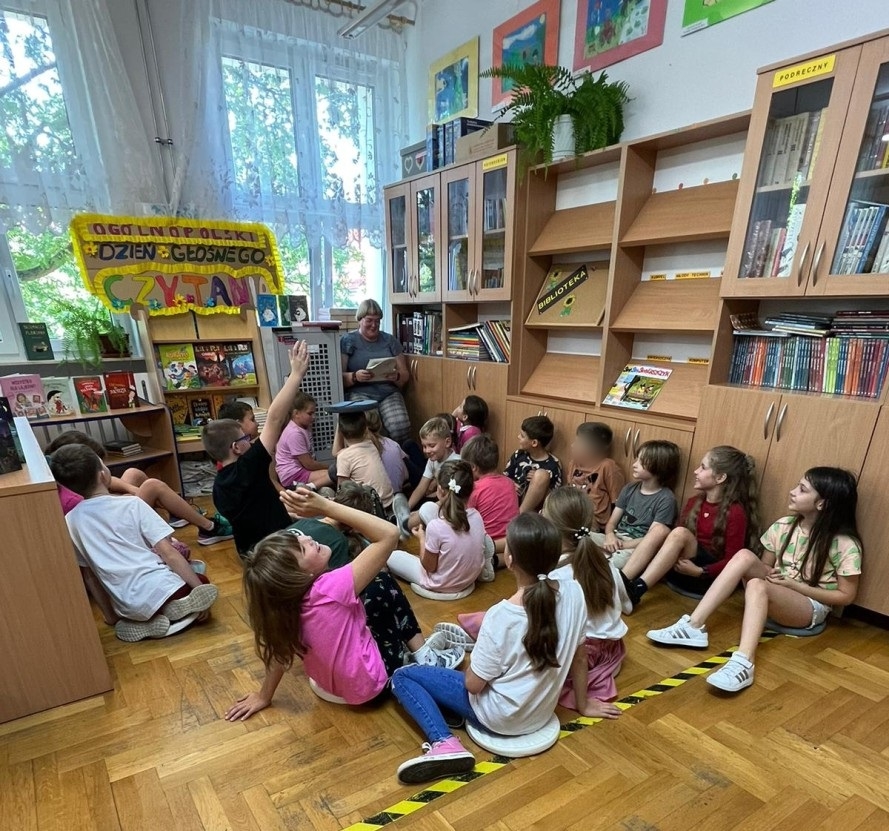 Dzieci siedzą na podłodze w bibliotece w siadzie skrzyżnym. Patrzą na stojącą przed nimi bibliotekarkę czytającą im tekst. 2 osoby podnoszą rękę do góry. W tle widać półki z książkami i kolorową planszę z napisem Ogólnopolski Dzień Głośnego Czytania