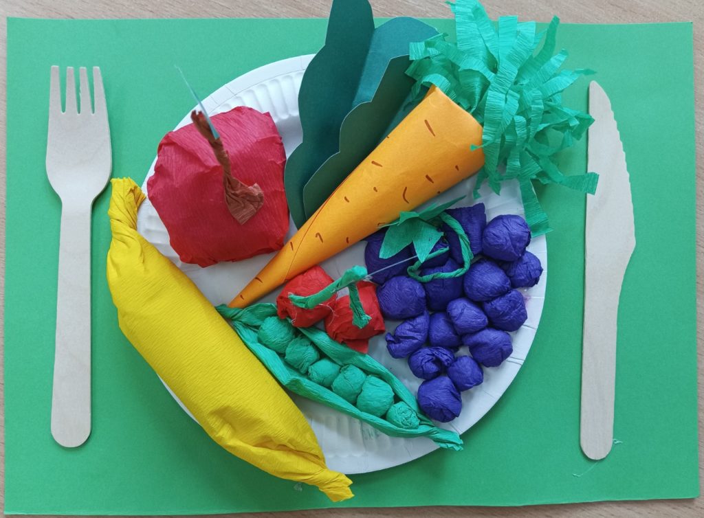 Zdjęcie przedstawia prace przestrzenną: na talerzyku papierowym przyklejone owoce i warzywa wykonane z kolorowego papieru i bibuły. Po lewej stronie talerza leży widelec, a po prawej nóż.