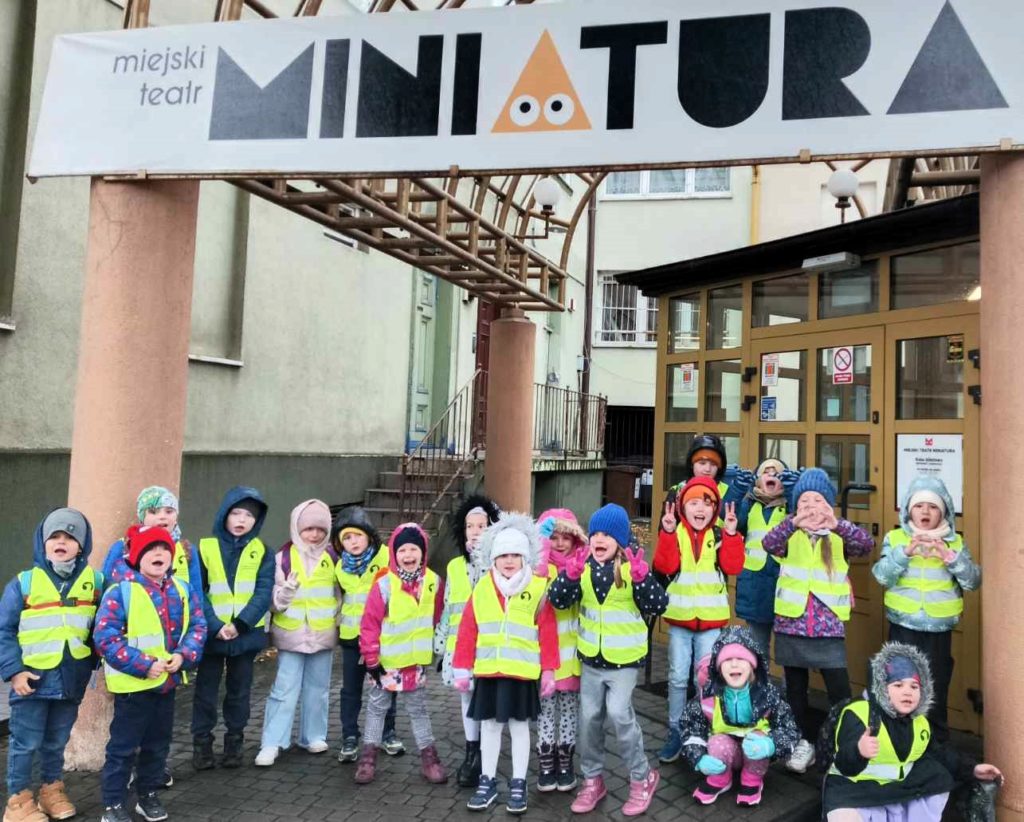 Grupa około 20 dzieci z uśmiechem na twarzy stoi przed wejściem do Teatru Miniatura.