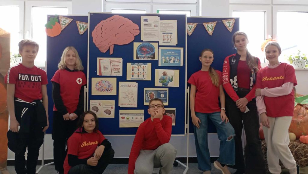 Na półpiętrze szkolnego korytarza na tle tablicy z plakatem na temat udaru mózgu stoją lub kucają uczniowie Koła PP HOPR w czerwonych koszulkach pozując do zdjęcia.