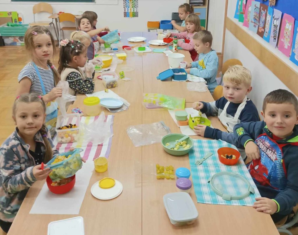 W klasie przy stolikach, które są połączone po 2 siedzą dzieci. Na stolikach jest dużo pojemników i produktów potrzebnych do zrobienia sałatek owocowych.