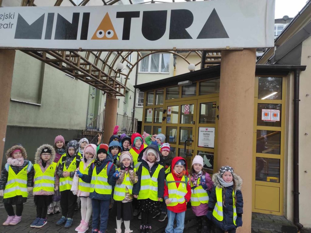 Grupa około 20 dzieci z uśmiechem na twarzy stoi przed wejściem do Teatru Miniatura.