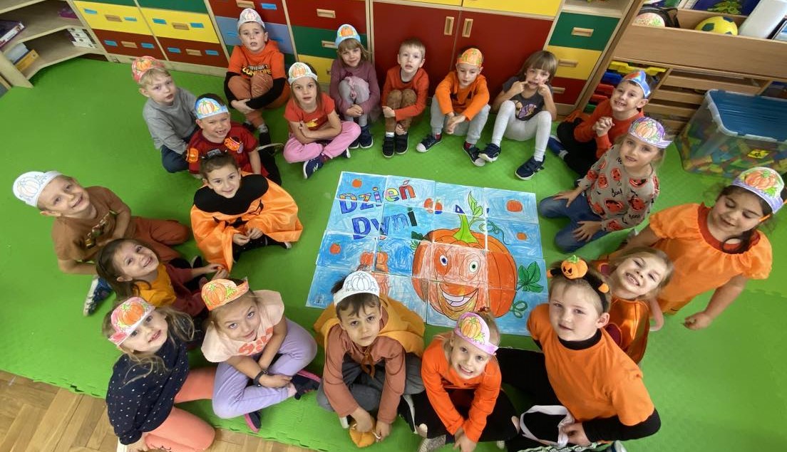  ok. 20 dzieci ubrana na pomarańczowo, niektórzy mają na głowie opaski z papierową dynią. Dzieci siedzą w kole na macie. W środku leży kolorowanka wielkformatowa.