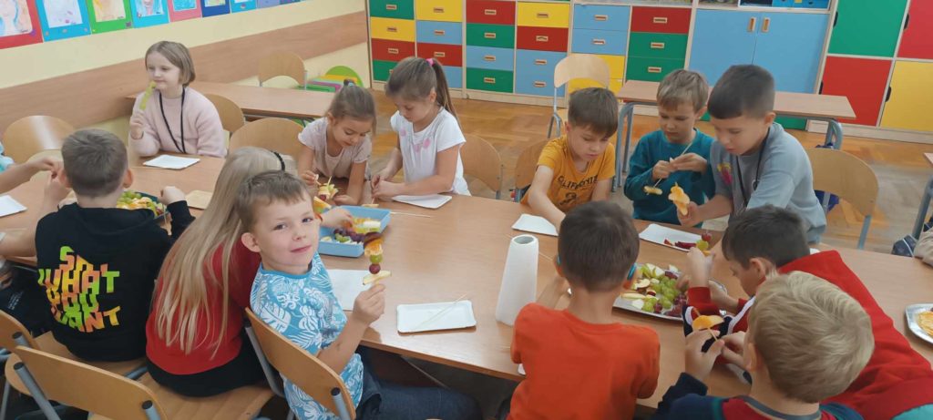 Dzieci w klasie szkolnej siedzące przy długim stole na którym przygotowują szaszłyki z kolorowych owoców.