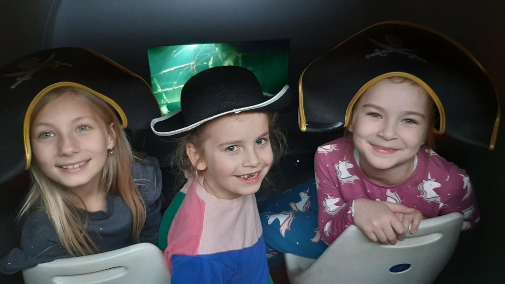 Trzy uśmiechnięte dziewczynki w pirackich czapkach na głowach siedzą w batyskafie. Za nimi monitor, na którym wyświetla się film przedstawiający wrak statku pod wodą.