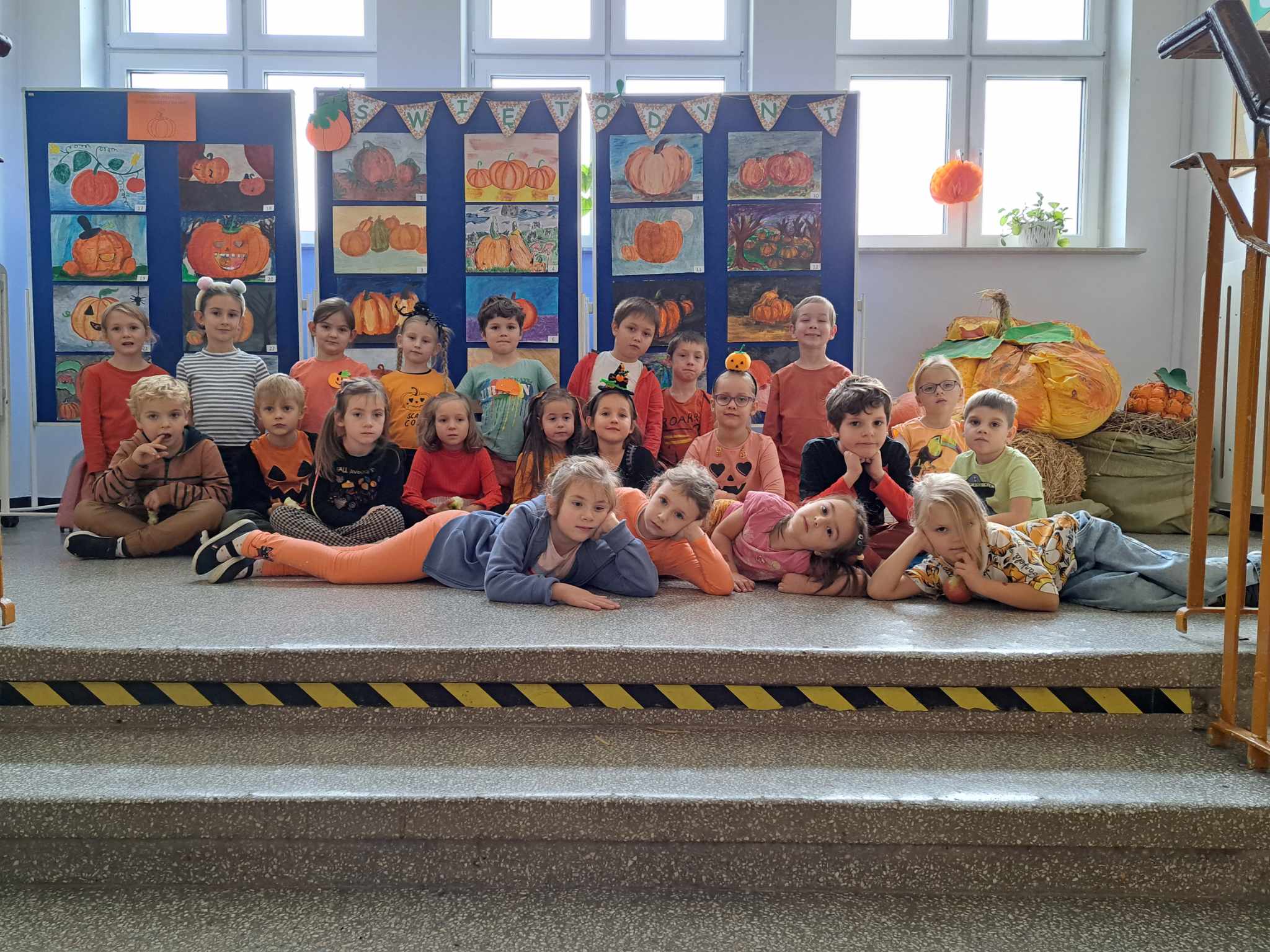 Ok. 20 dzieci ubrana na pomarańczowo kuca na korytarzu szkolnym. W tle prace plastyczne z motywem dyni oraz napis Dzień dyni.