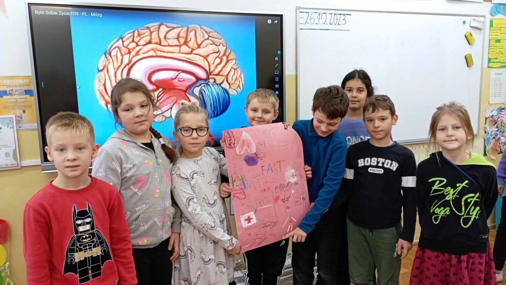 W sali lekcyjnej na tle tablicy multimedialnej wyświetlającej obraz ludzkiego mózgu stoi grupa 8 uczniów klasy 2c. Troje z nich trzyma plakat na temat udaru mózgu.