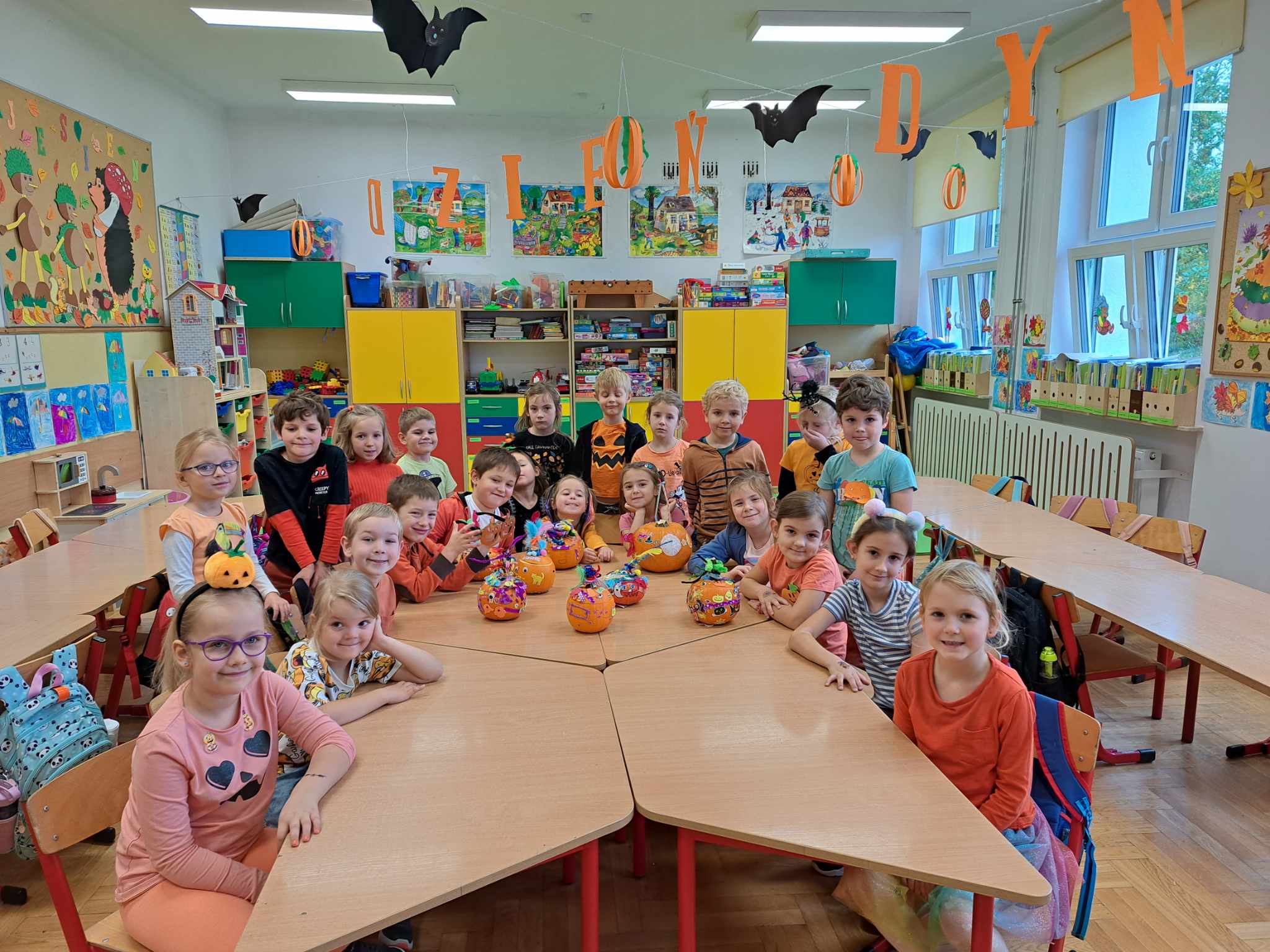 Ok. 20 dzieci ubrana na pomarańczowo stoi lub siedzi przy stolikach, na których znajdują się własnoręcznie wykonane dynie. 