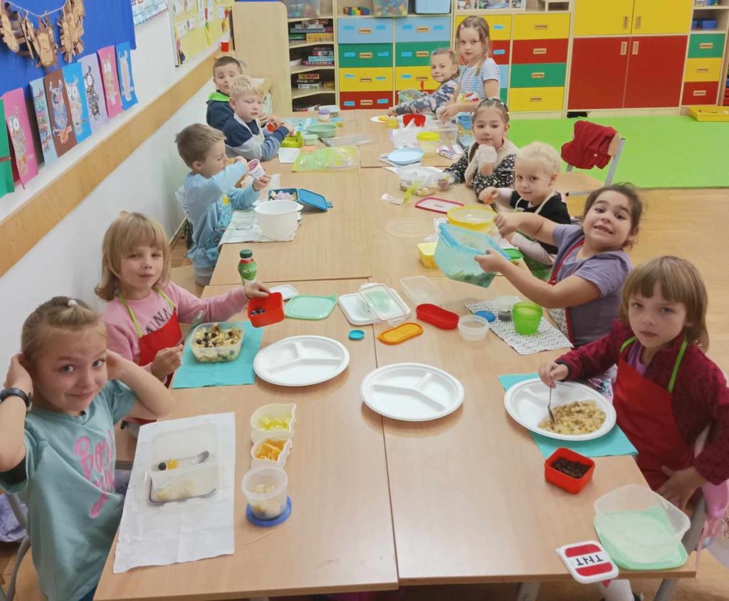 W klasie przy stolikach, które są połączone po 2 siedzą dzieci. Na stolikach jest dużo pojemników i produktów potrzebnych do zrobienia sałatek owocowych.