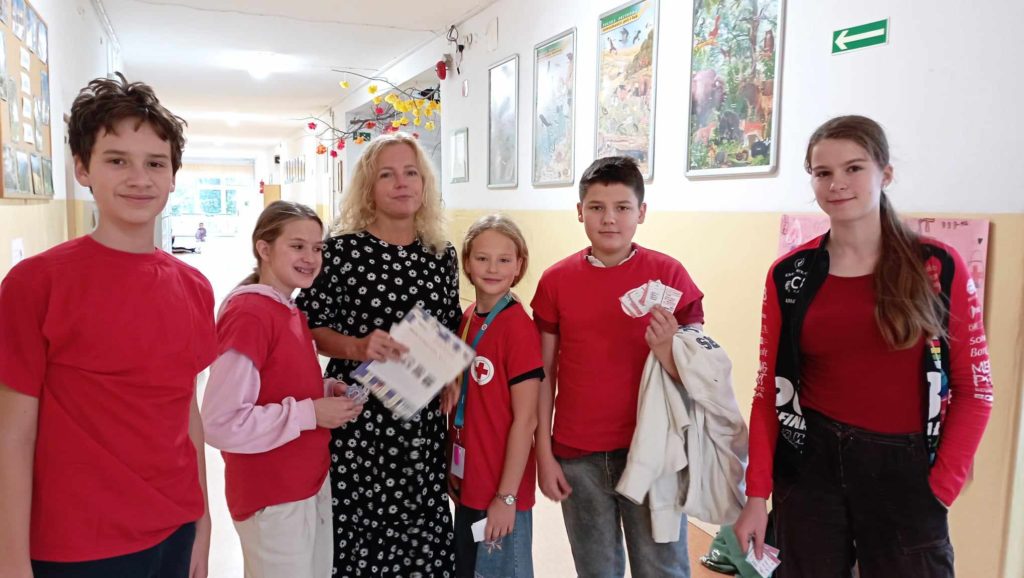 Na szkolnym korytarzu stoi nauczycielka trzymająca kartki. W koło niej 5 uczniów Koła PP HOPR w czerwonych koszulkach trzymających ulotki na temat udaru mózgu.