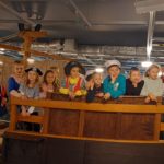 Grupa zadowolonych dzieci ustawiona w rzędzie na pokładzie atrapy żaglowca. Co drugi uczeń przebrany w pirata lub marynarza. Po lewej stronie za dziećmi jest opuszczony żagiel, a na dole na burcie żaglowca wisi tablica poglądowa z olinowaniem żaglowca.