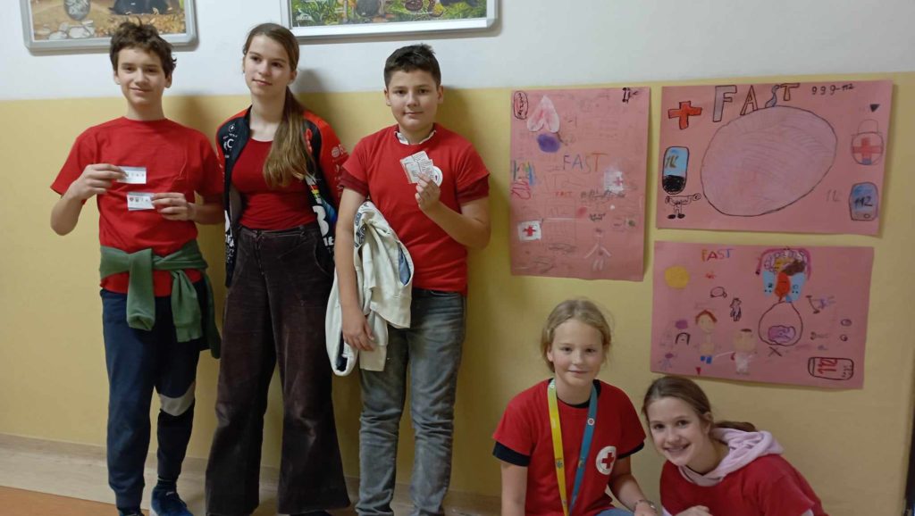 Na szkolnym korytarzu, na tle ściany z plakatami na temat udaru mózgu stoi lub kuca 5 uczniów Koła PP HOPR w czerwonych koszulkach trzymający w dłoniach ulotki na temat udaru mózgu.