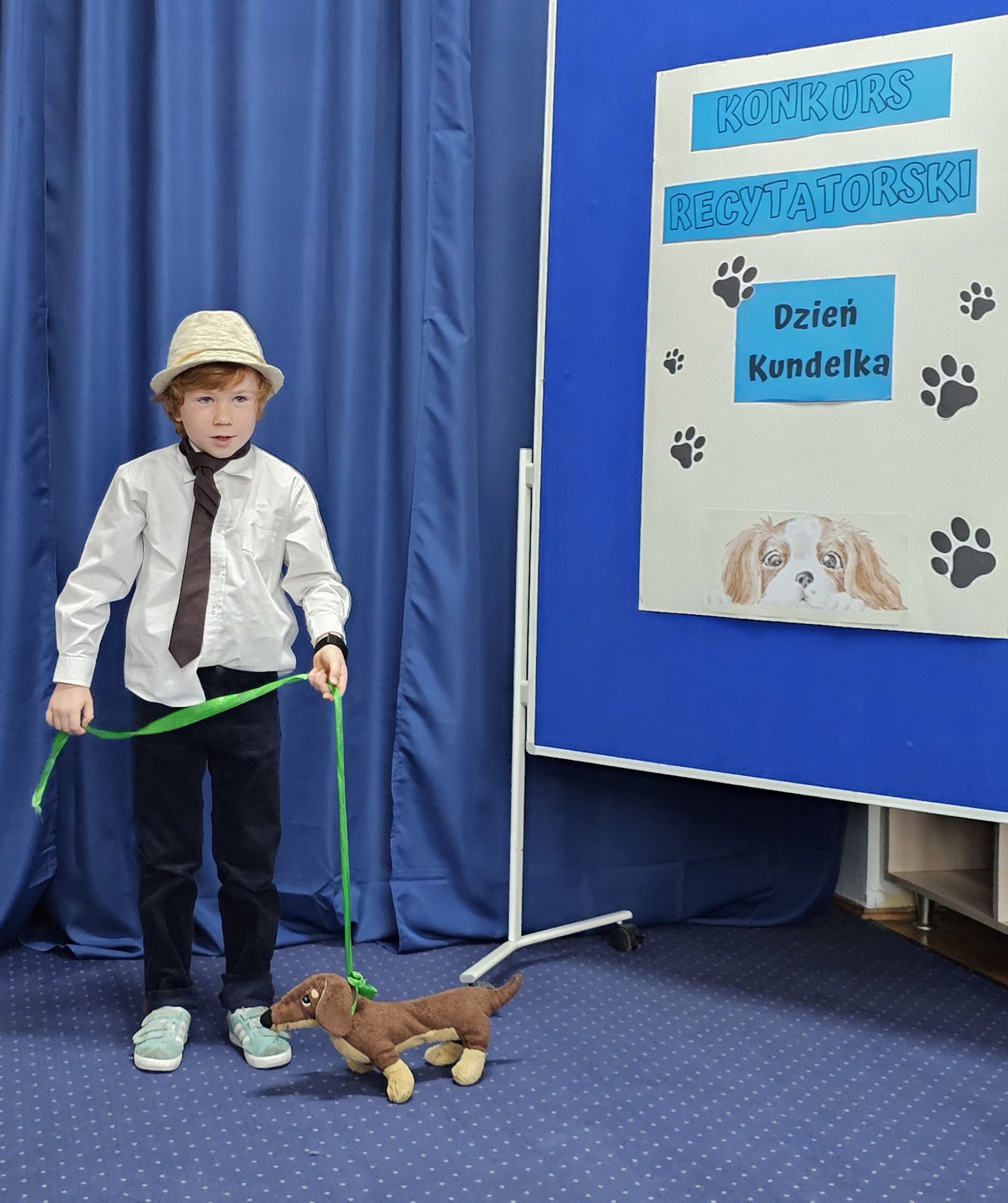 W sali stoi chłopiec w kapeluszu. W ręku trzyma smycz z przywiązaną maskotką psa. Z boku widać tablicę z napisem: konkurs recytatorski, dzień kundelka