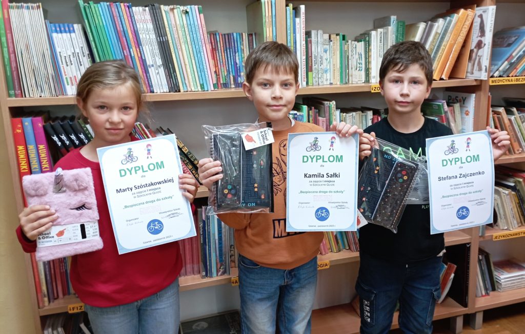 Dwaj chłopcy i jedna dziewczynka pozują do zdjęcia z dyplomem oraz nagrodą w rękach. W tle na regałach stoją książki.