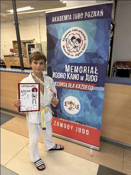 Chłopiec ubrany w judogę stoi na tle plakatu i trzyma dyplom za zajęcie 3 miejsca w zawodach międzynarodowych