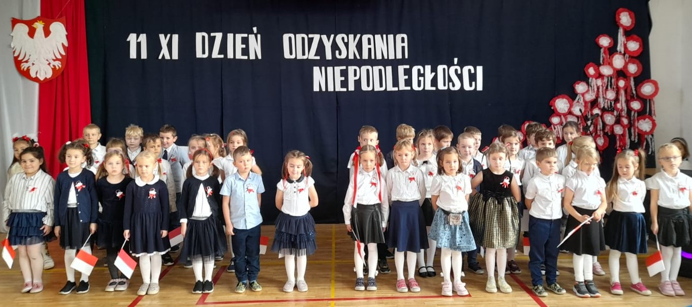 ponad 30 dzieci ubranych na galowo stoi na sali na baczność. Niektóre dzieci mają w ręku małe flagi Polski. W tyle godło Polski na tle biało czerwonej flagi, a nad dziećmi napis: 11 XI dzień odzyskania niepodległości