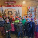 Ok. 20 dzieci stoi pozuje z św. Mikołajem. W tle dekoracje świąteczne oraz napis „PRZYWIDZKA OSADA SW. MIKOLAJA".