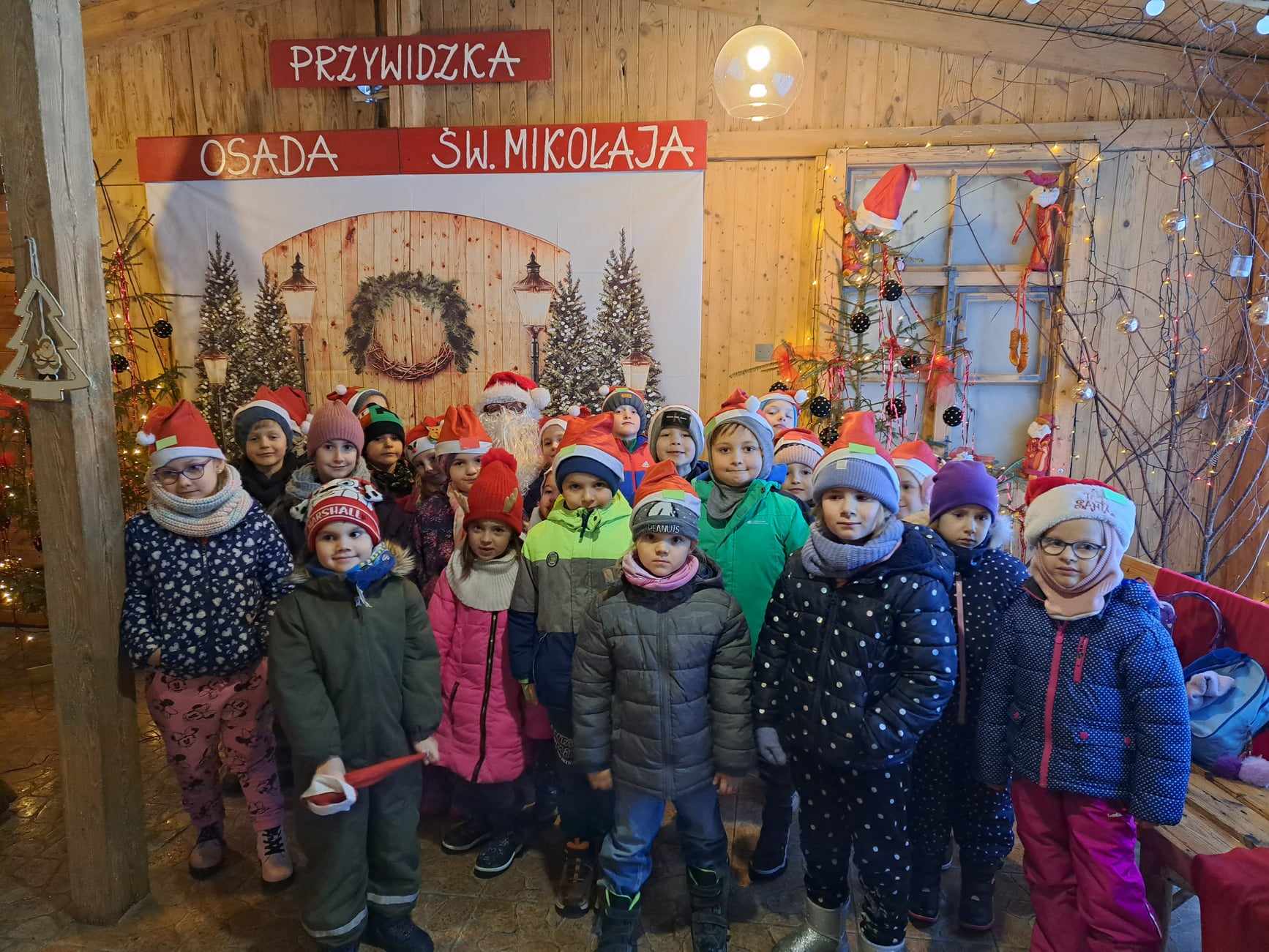 Ok. 20 dzieci stoi pozuje z św. Mikołajem. W tle dekoracje świąteczne oraz napis „PRZYWIDZKA OSADA SW. MIKOLAJA".