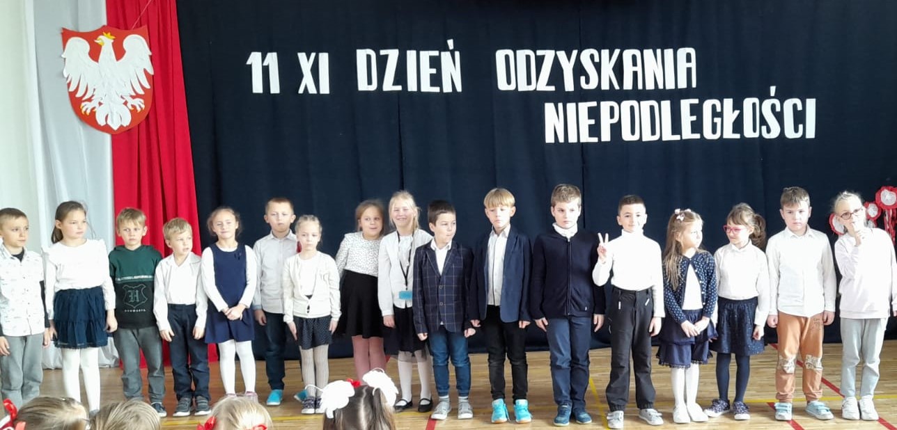 17 ubranych na galowo dzieci stoi na sali. W tyle godło Polski na tle biało czerwonej flagi, a nad dziećmi napis: 11 XI dzień odzyskania niepodległości