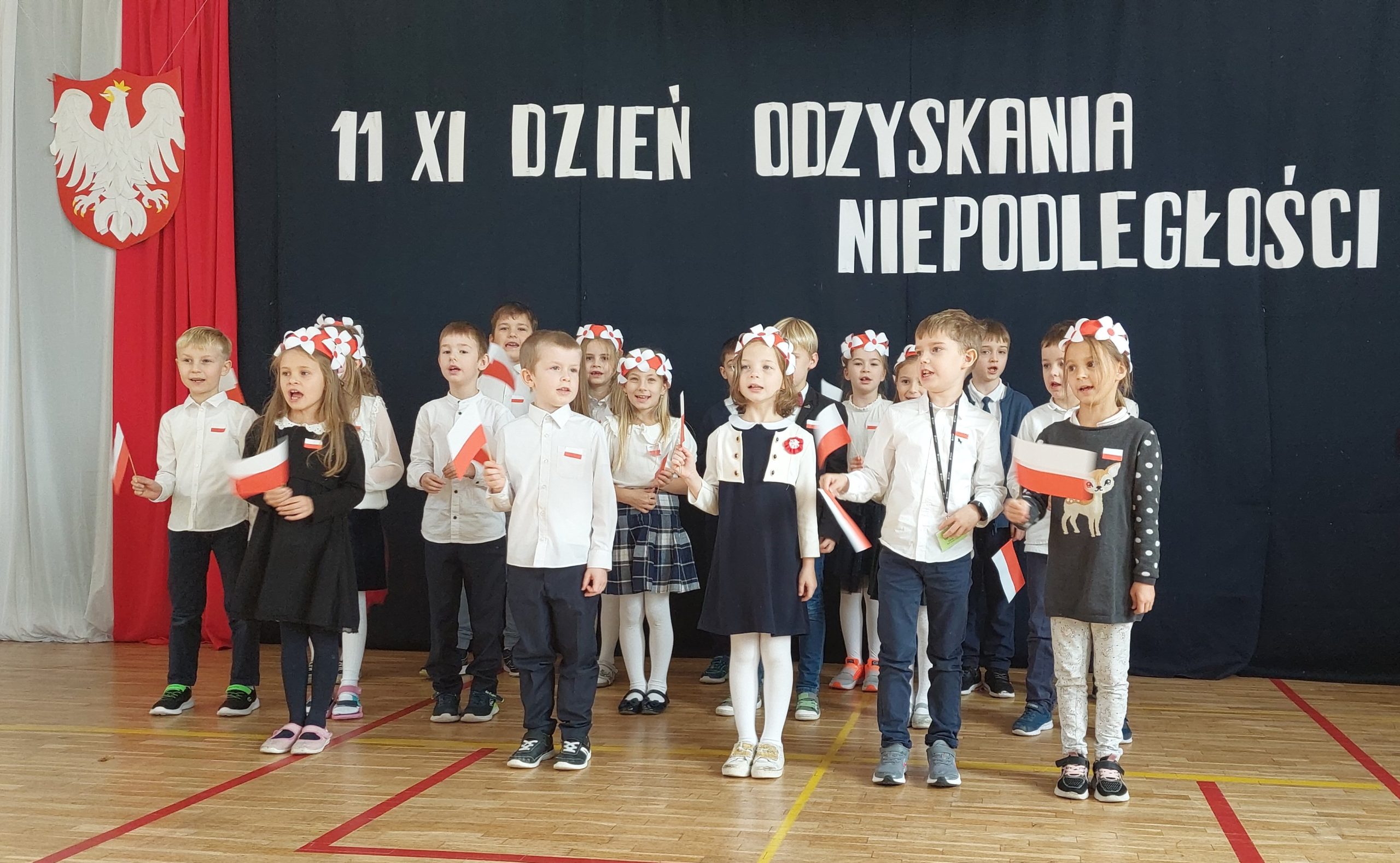 17 ubranych na galowo dzieci stoi na sali, w ręku trzymają białe i czerwone wstążki. W tyle godło Polski na tle biało czerwonej flagi, a nad dziećmi napis: 11 XI dzień odzyskania niepodległości