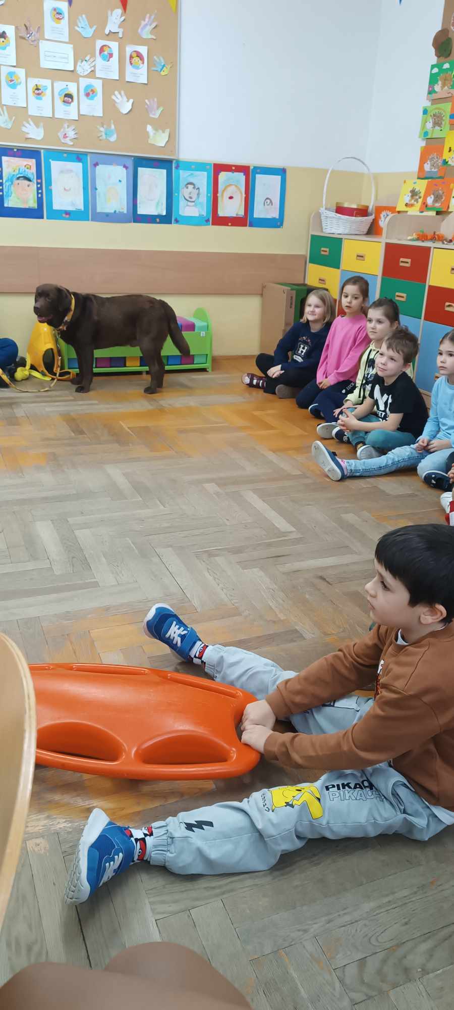 Klasa szkolna, na pierwszym planie siedzący chłopiec na podłodze trzyma pomarańczową deskę ratunkową. Po prawej stronie grupa dzieci obserwująca chłopca. Na wprost stoi duży brązowy pies.