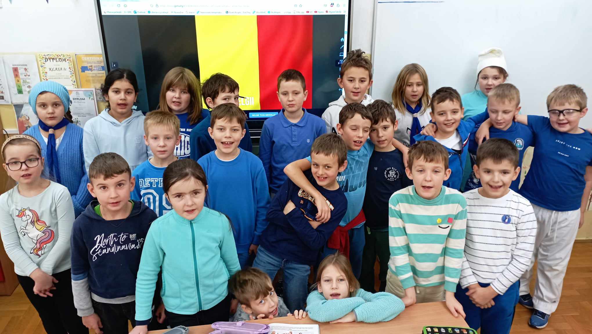 23 uczniów klasy 2c przebranych za Smerfy stoi lub kuca w sali na tle tablicy multimedialnej wyświetlającej flagę Belgii.