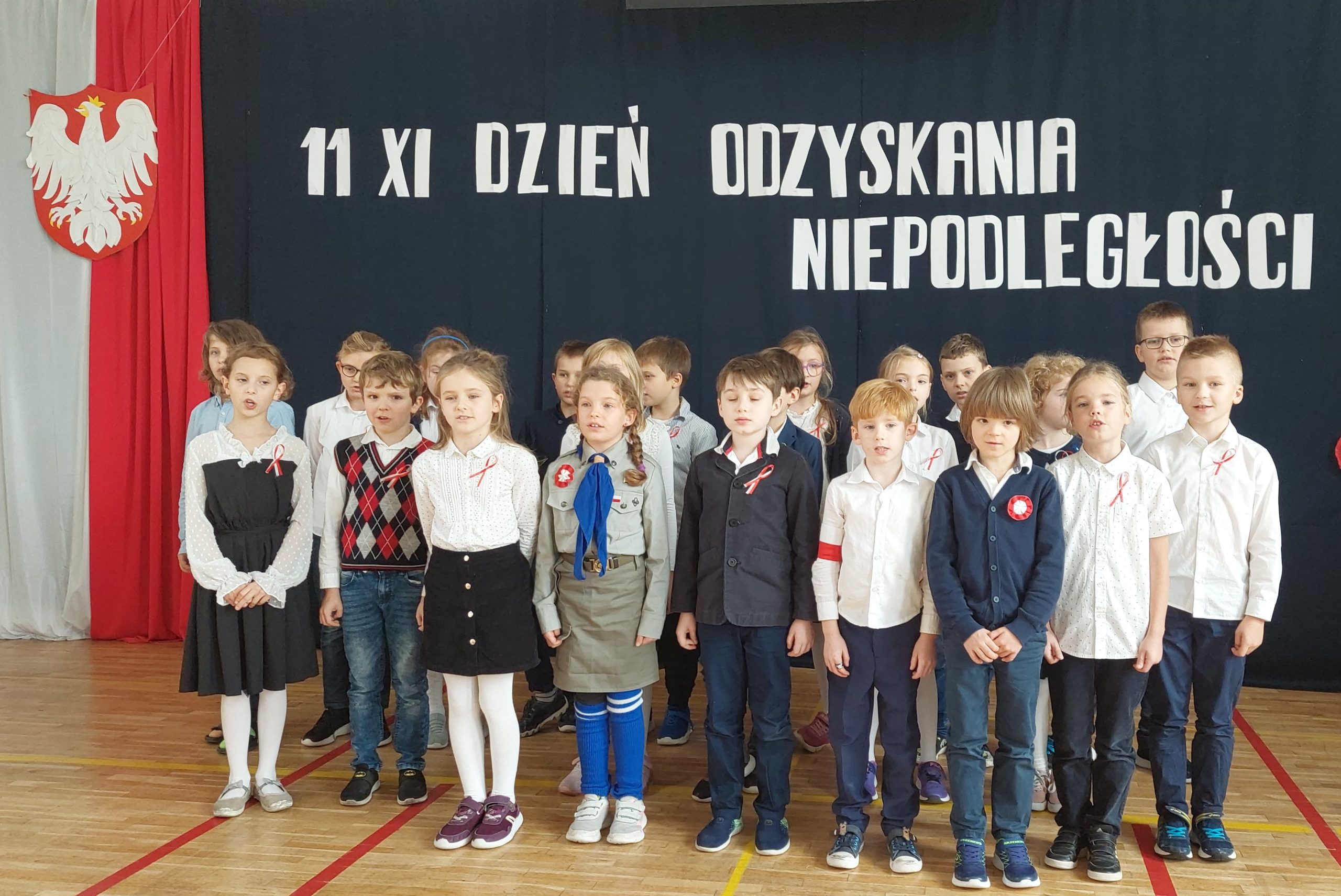 Ponad 20 ubranych na galowo dzieci stoi na sali. W tyle godło Polski na tle biało czerwonej flagi, a nad dziećmi napis: 11 XI dzień odzyskania niepodległości