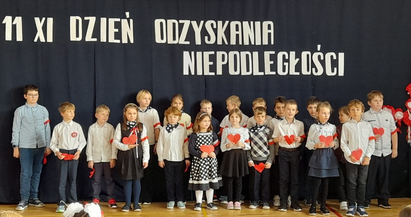 20 ubranych na galowo dzieci stoi na sali. W rękach trzymają czerwone papierowe serca. W tyle godło Polski na tle biało czerwonej flagi, a nad dziećmi napis: 11 XI dzień odzyskania niepodległości