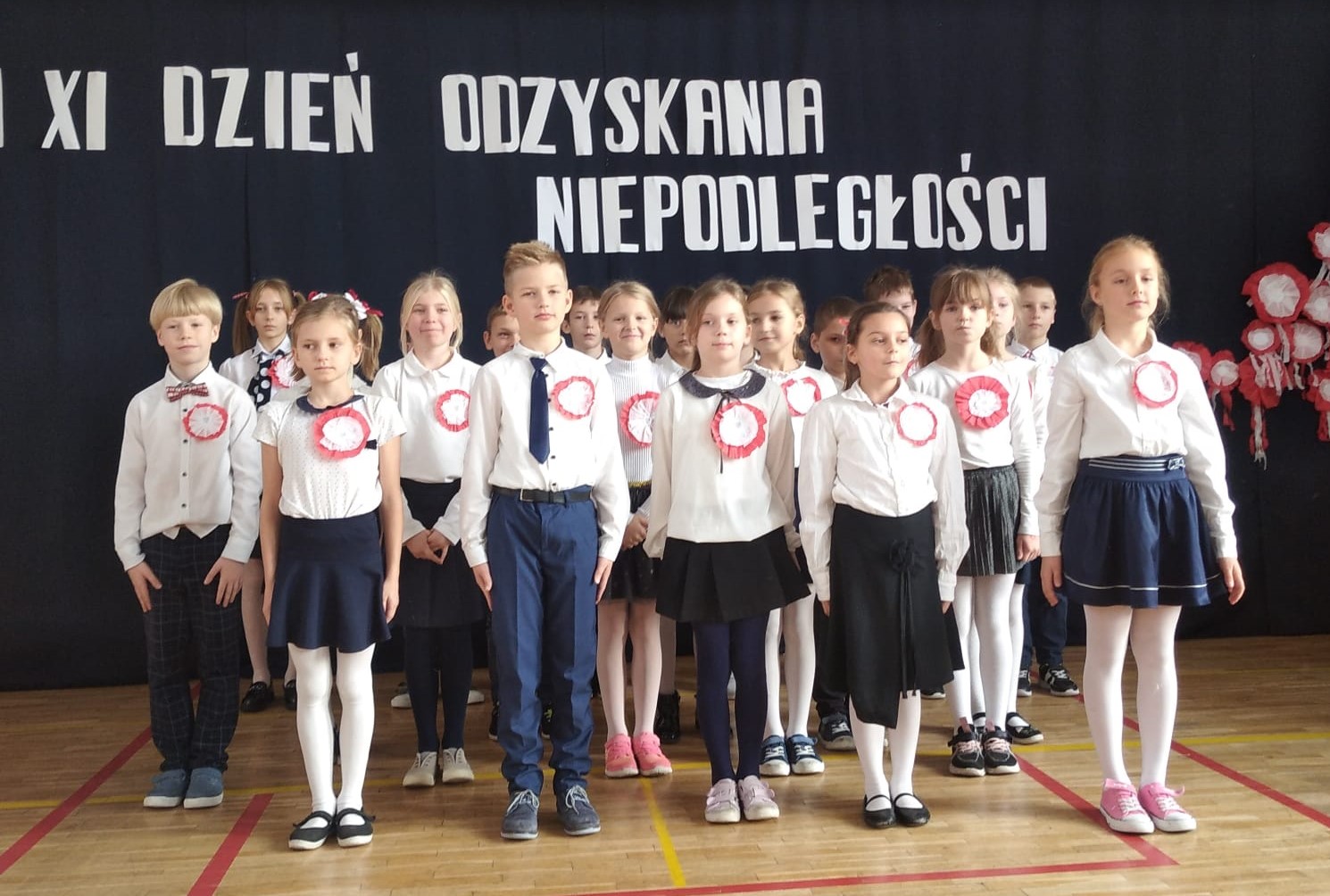 18 ubranych na galowo dzieci stoi na sali. W tyle godło Polski na tle biało czerwonej flagi, a nad dziećmi napis: 11 XI dzień odzyskania niepodległości