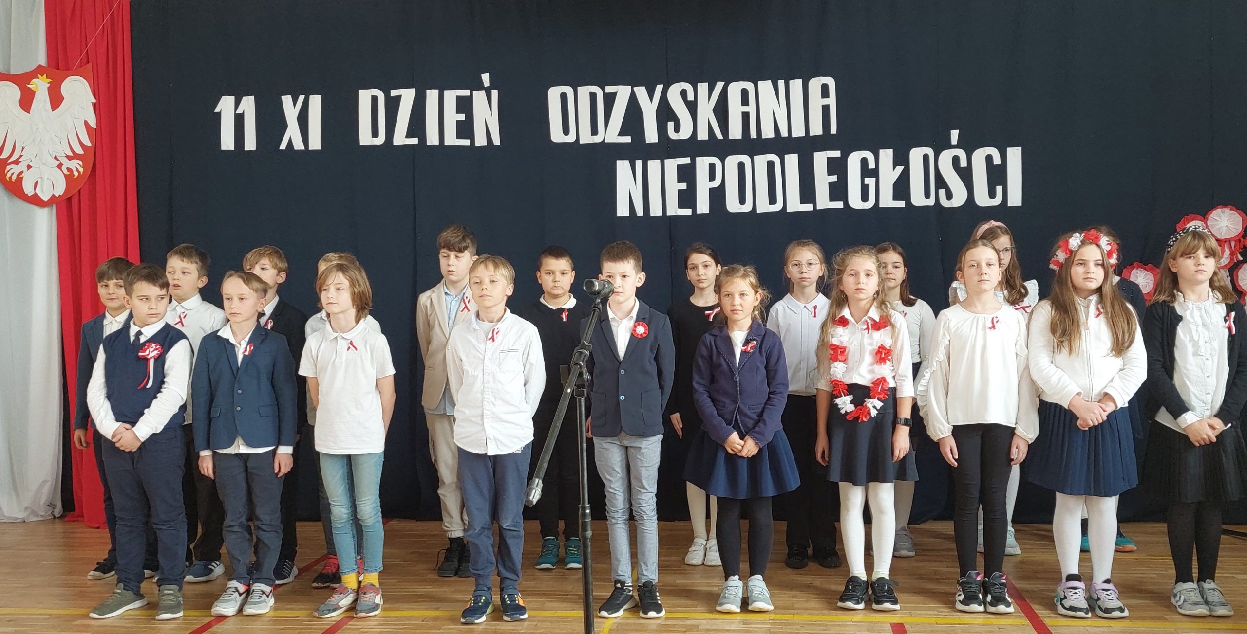 Ponad 20 ubranych na galowo dzieci stoi na sali. W tyle godło Polski na tle biało czerwonej flagi, a nad dziećmi napis: 11 XI dzień odzyskania niepodległości
