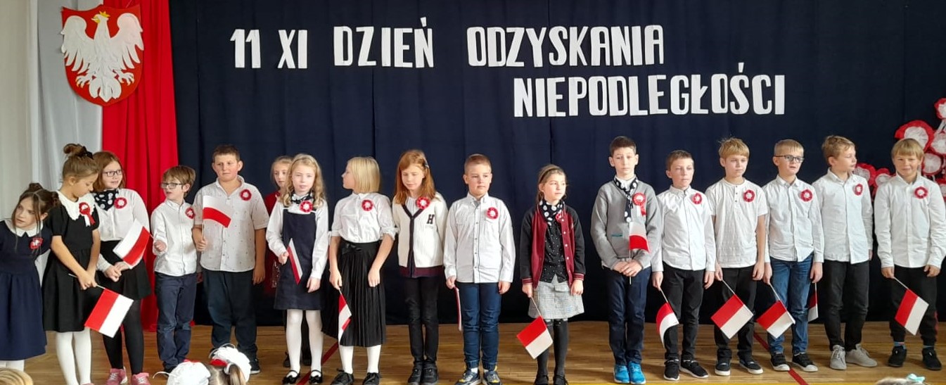 17 ubranych na galowo dzieci stoi na sali. Niektóre dzieci mają w ręku małe flagi Polski. W tyle godło Polski na tle biało czerwonej flagi, a nad dziećmi napis: 11 XI dzień odzyskania niepodległości