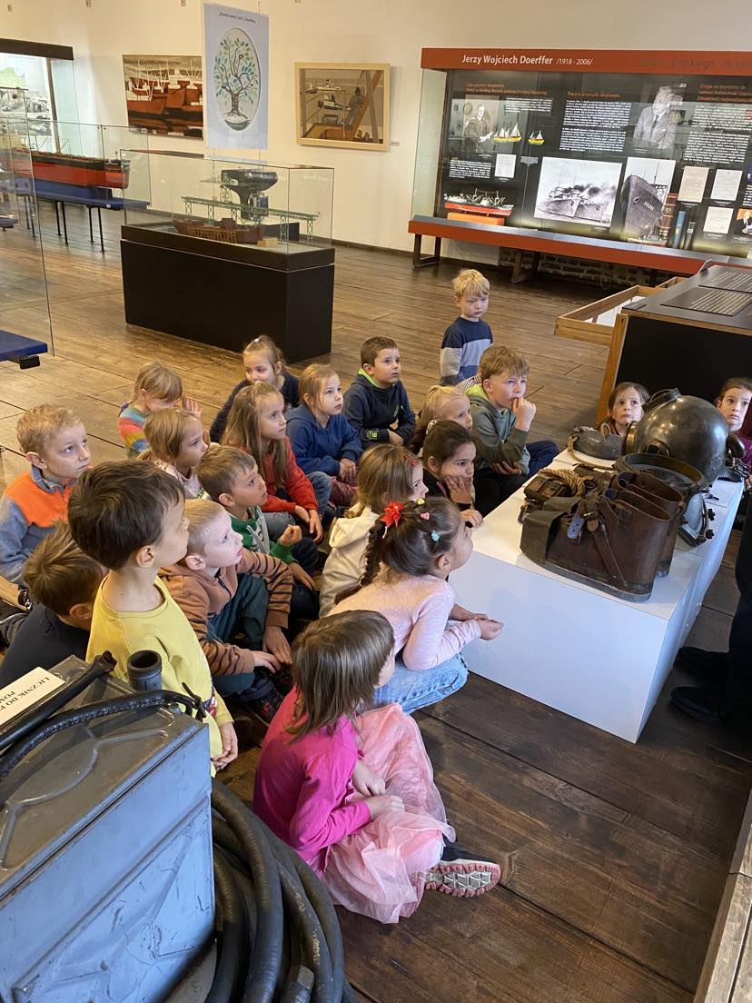 ok. 20 dzieci siedzi na podłodze w sali z eksponatami z dziedziny archeologii podwodnej.
