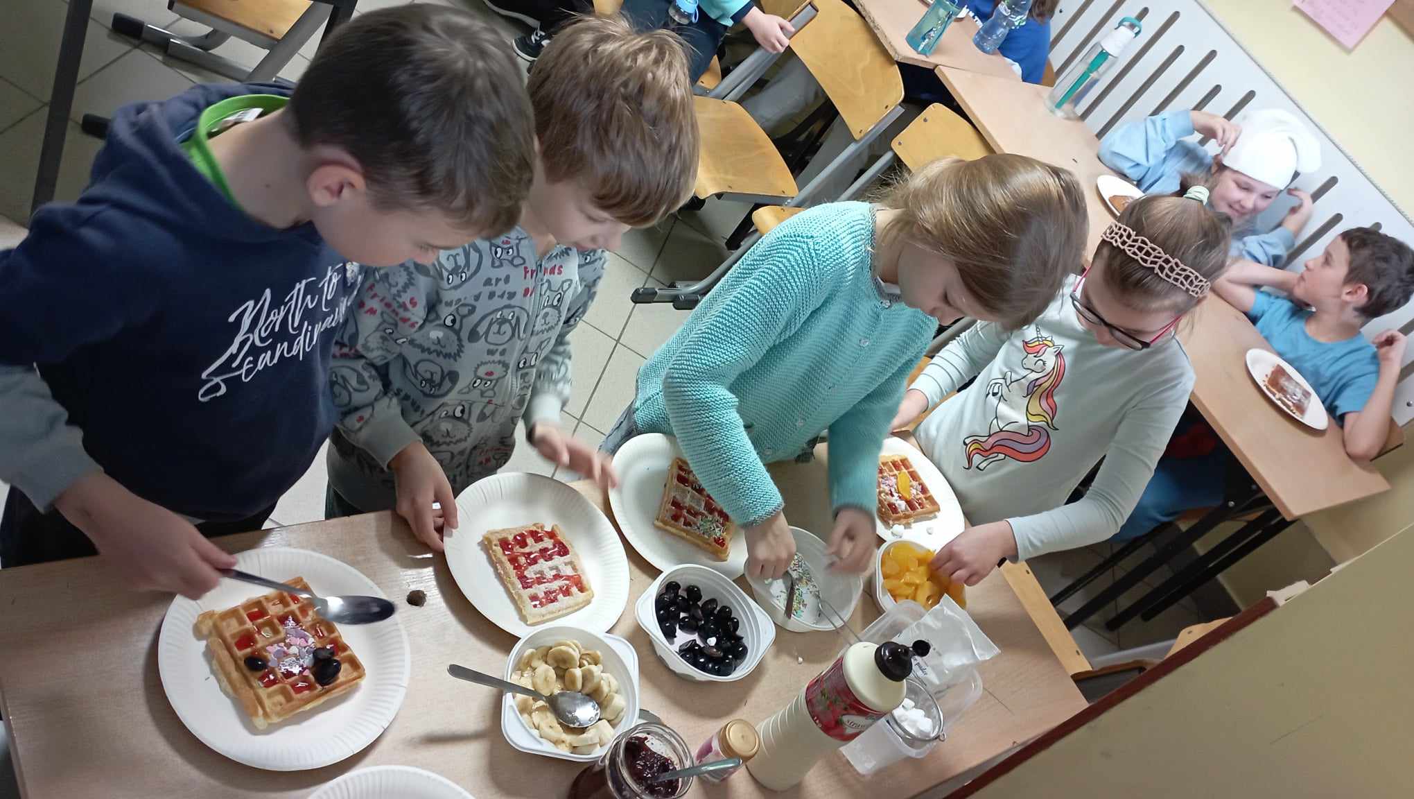 4 uczniów klasy 2c stoi przy stoliku z produktami spożywczymi i nakłada sobie smakołyki na swojego gofra. W tle uczniowie siedzący przy stole z talerzykami i goframi.