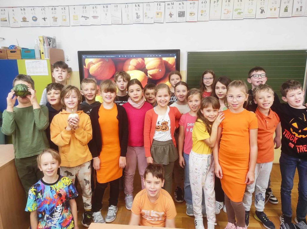 Grupa uczniów stoi w klasie przed tablicą multimedialną na której wyświetlone są dynie. Większość uczniów ubrana jest na pomarańczowo. Dwóch chłopców trzyma w ręku małą dynię.