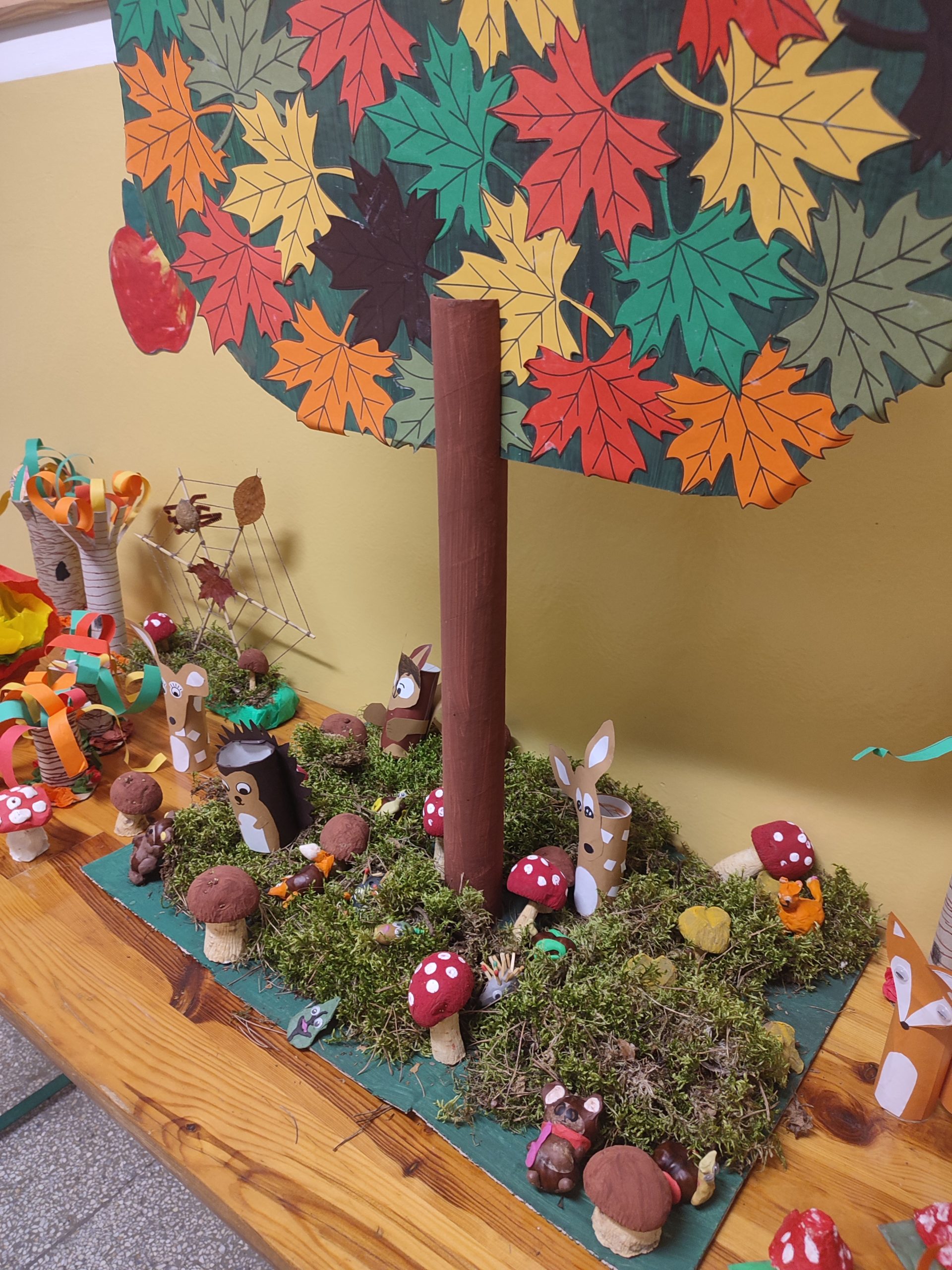 Jesienna wystawa prac plastycznych i technicznych. Na stole stoi wysokie drzewo z tektury a w koło niego pośród mchu rozstawione zwierzątka, grzyby i drzewa z papierowych rolek, plasteliny i warzyw.
