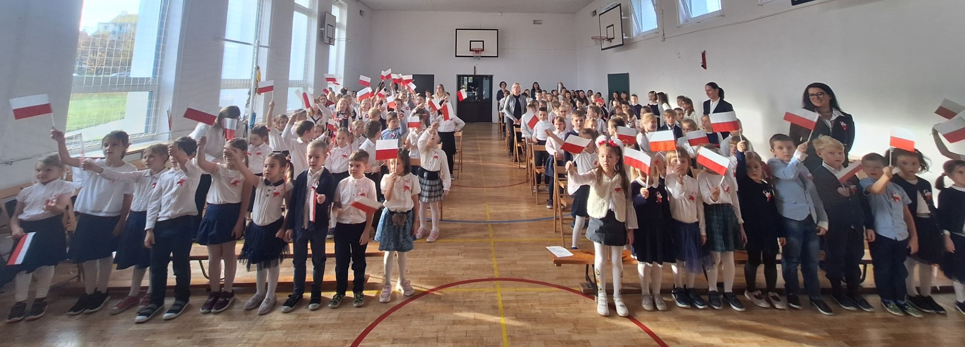 Na sali gimnastycznej stoi w rzędach kilkadziesiąt ubranych na galowo dzieci. Niektóre z nich trzymają podniesione w górę małe flagi Polski.