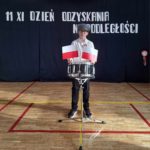 Na sali gimnastycznej stoi chłopiec w harcerskiej czapce, przed stoi werbel ustrojony 2 małymi flagami Polski. Za Chłopcem w tle widać napis: „11 XI DZIEŃ ODZYSKANIA NIEPODLEGŁOŚCI”