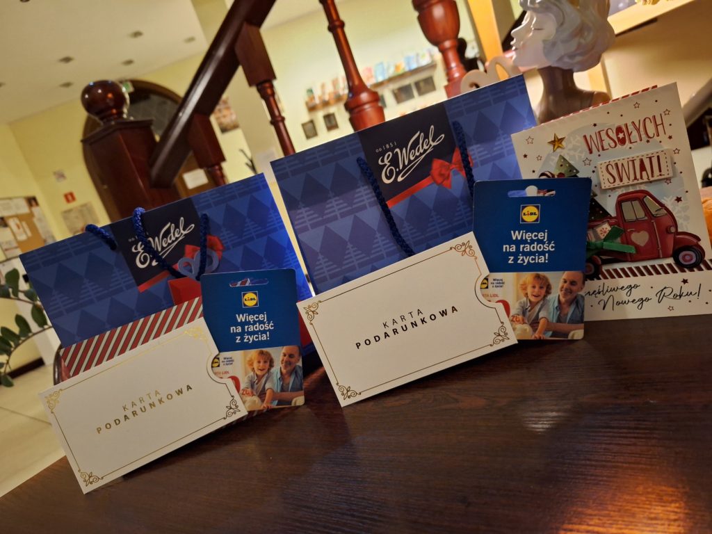Na stole stoją 2 torebki prezentowe i 2 koperty z kartami podarunkowymi oraz kartka świąteczna z napisem wesołych świąt.