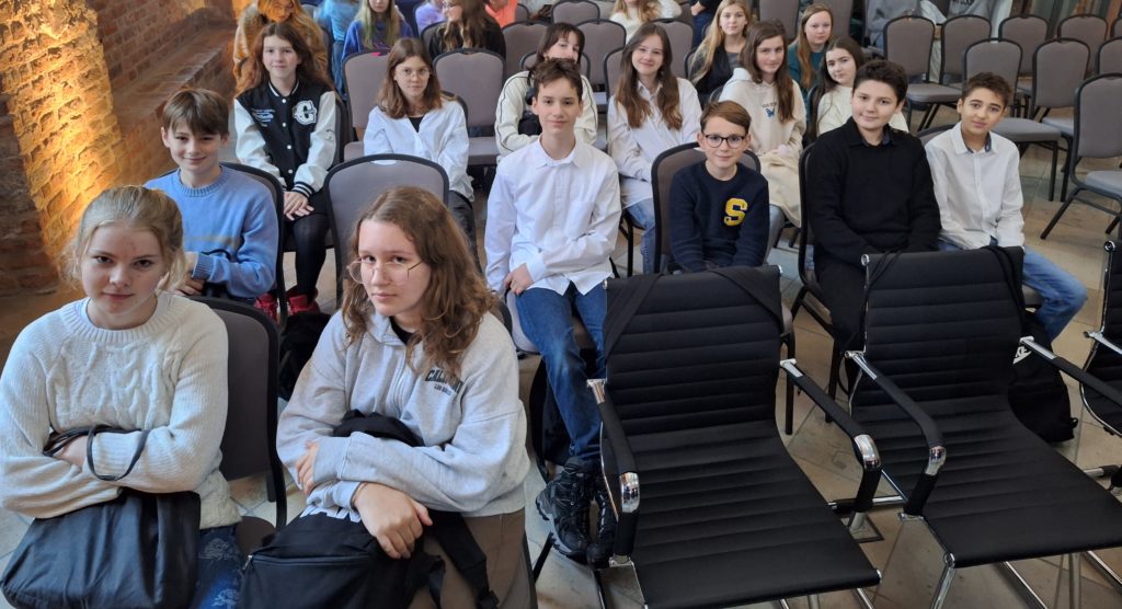 Grupa dzieci ubrana elegancko siedzi na krzesłach w sali konferencyjnej.